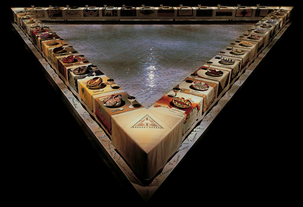 Œuvre d'art The Diner Party réalisée par Judy Chicago. Trois tables sont disposées de façon à créer un triangle. A chacune des place, se trouve un set de table brodé avec le nom d'une femme puissante (fictive ou réelle) et une assiette décorée.