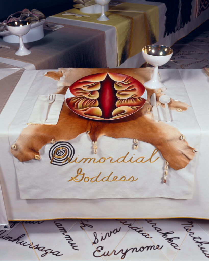 Zoom sur l'ouvre d'art de Judy Chicago. On est devant un set de table brodé "Primordial Goddess" et une assiette peinte (en rouge, jaune et orange). La forme peinte rappelle une fleur ou une vulve.